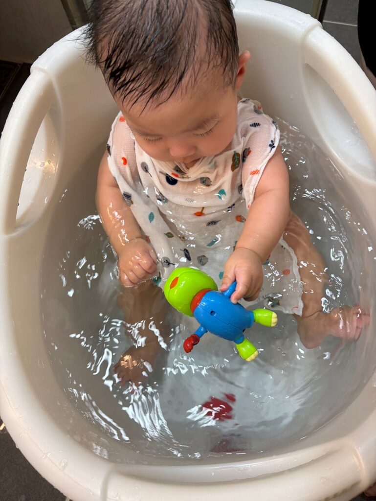 욕조에 앉은 6개월 아기가 수영하는 크롱 장난감에 관심을 보이는 모습
